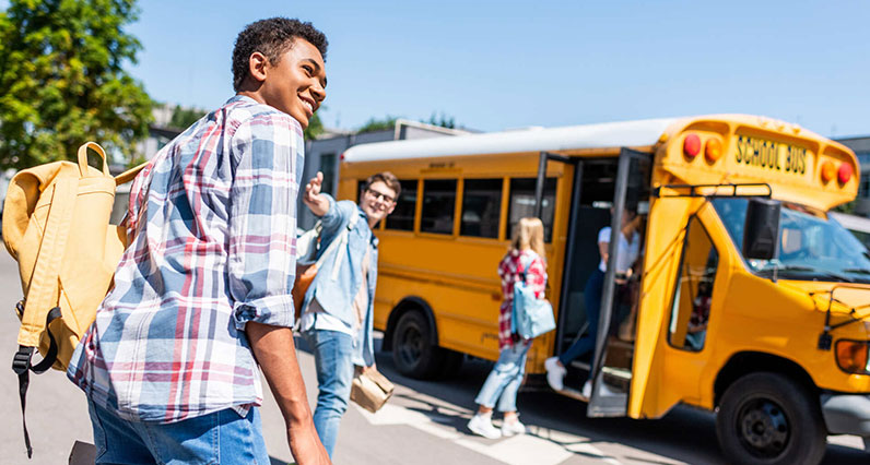 rear view of smiling teen african american schoolboy walking to school bus with his classmates
